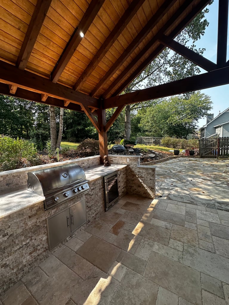 Outdoor patio with wooden pergola, built-in grill, stone countertop, and paver surface surrounded by landscaping and trees